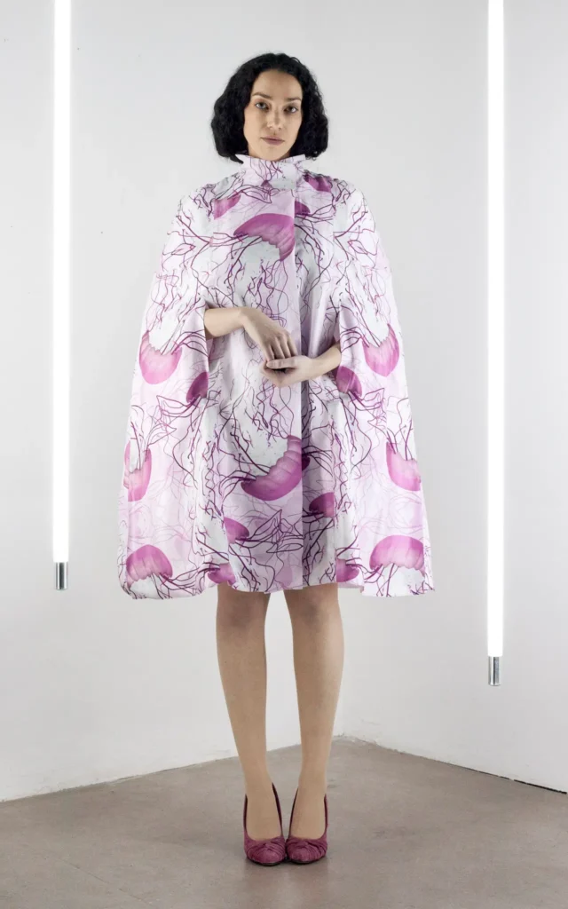 Model wearing a high-collar cape dress in pink, printed with jellyfish-inspired motifs.